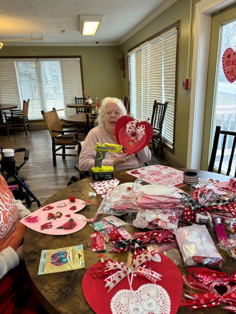 An elderly woman is sitting at a table decorating for valentine 's day.
