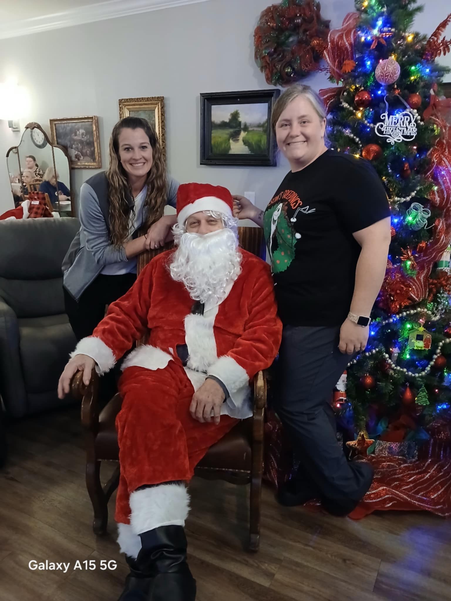 A man dressed as santa claus is sitting in a chair next to two women and a christmas tree.