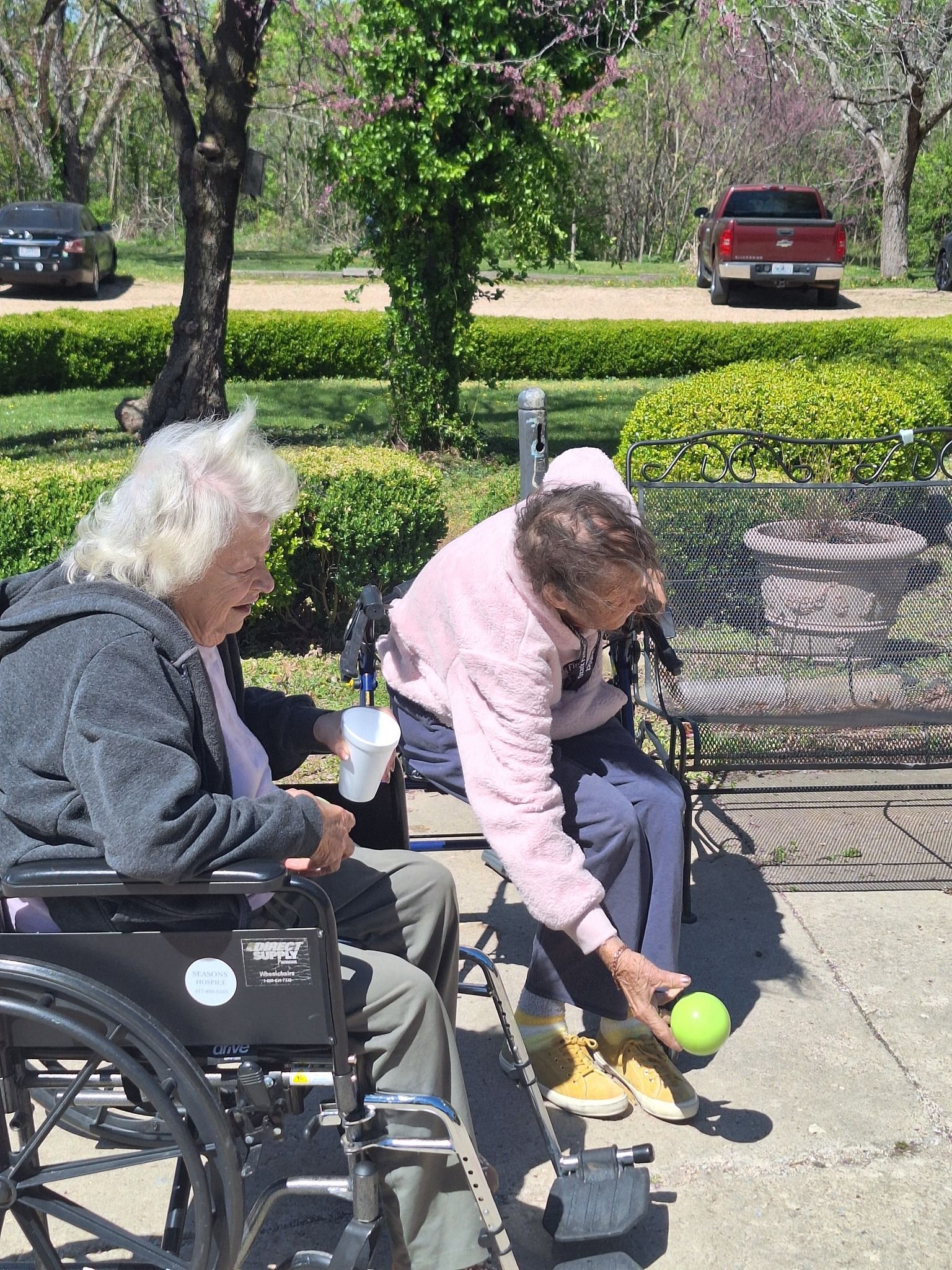 Two elderly people in wheelchairs are playing bocce ball in a park.