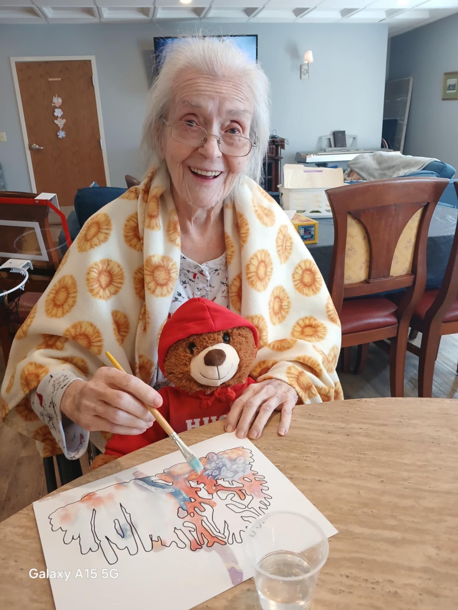 An elderly woman is sitting at a table painting with a teddy bear.