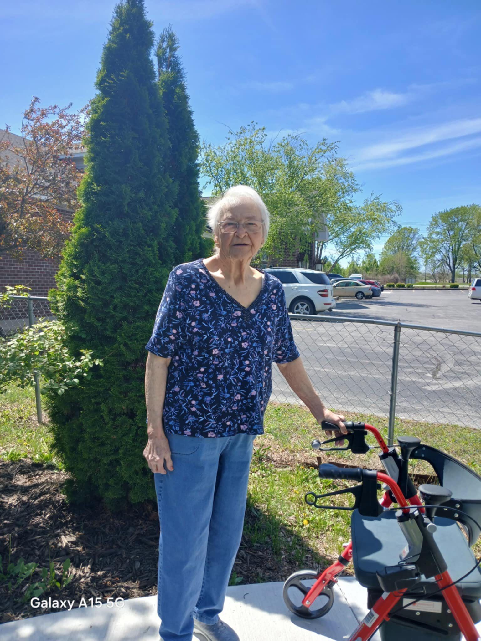 An elderly woman is standing next to a walker in a parking lot.