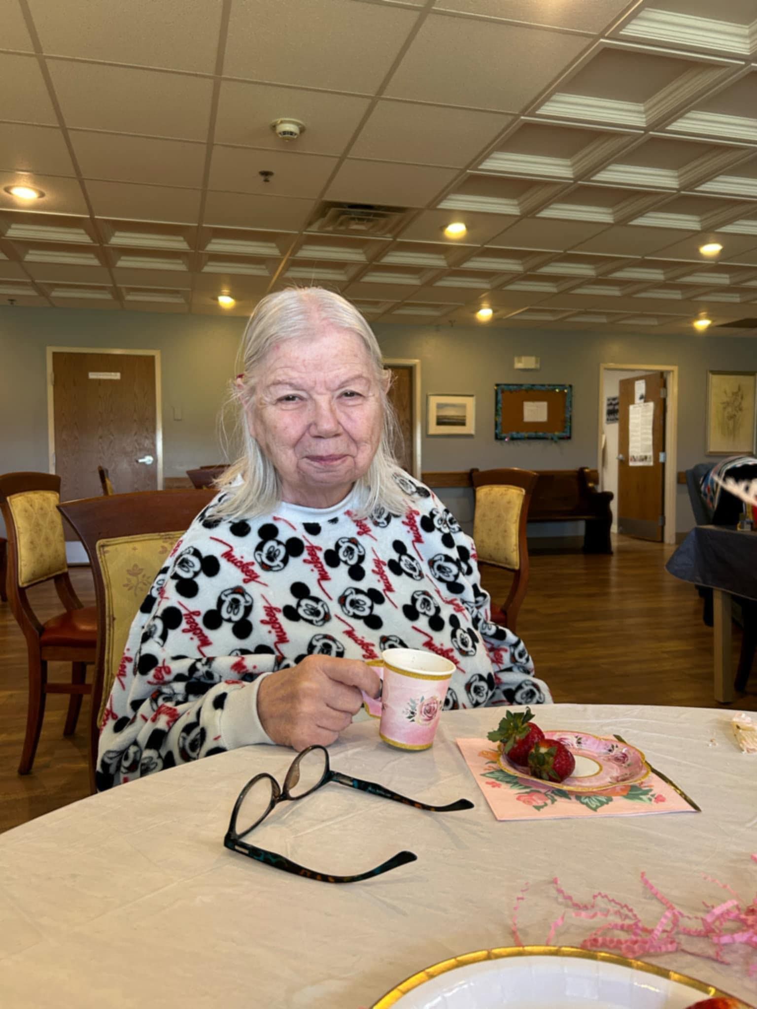 An elderly woman is sitting at a table drinking a cup of tea.