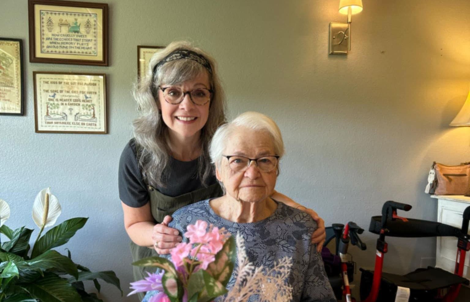 A woman is standing next to an older woman in a living room holding flowers.