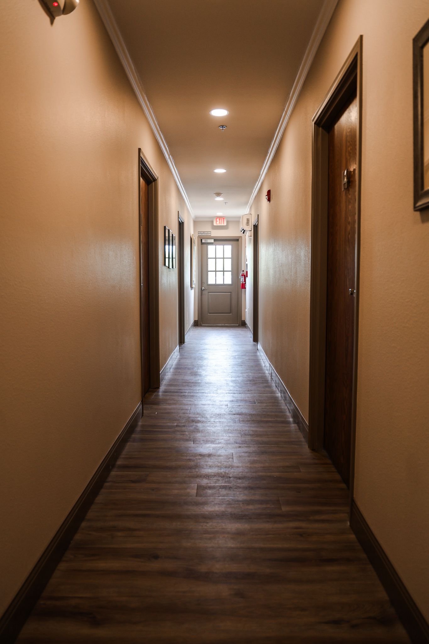 A long hallway with wooden floors and brown walls leading to a door.