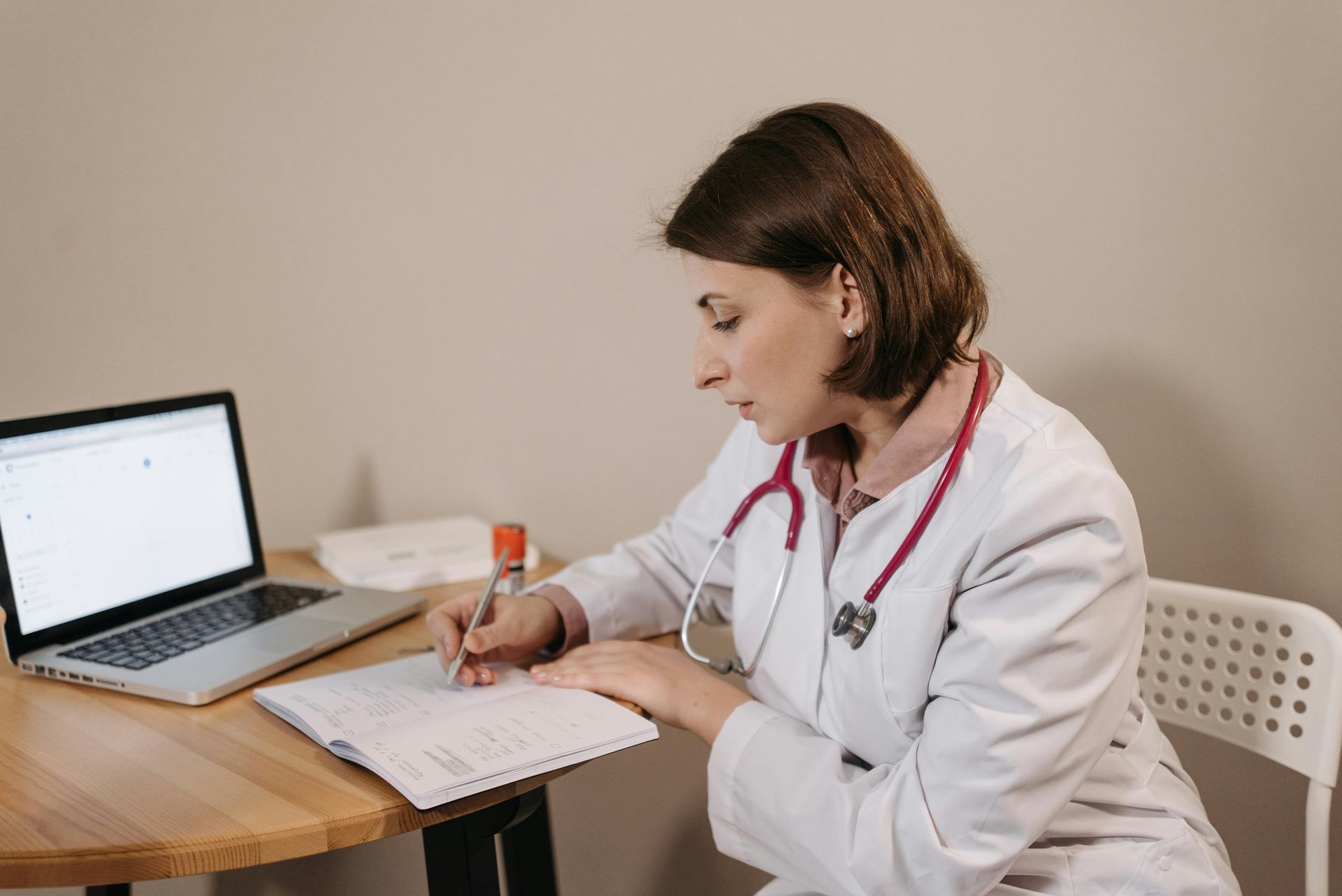 A female doctor is sitting at a table with a laptop and a notebook.