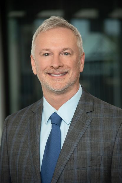 A man in a suit and blue shirt is smiling in front of a brick wall.