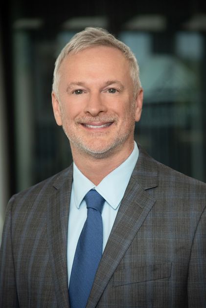 A man in a suit and blue shirt is smiling in front of a brick wall.