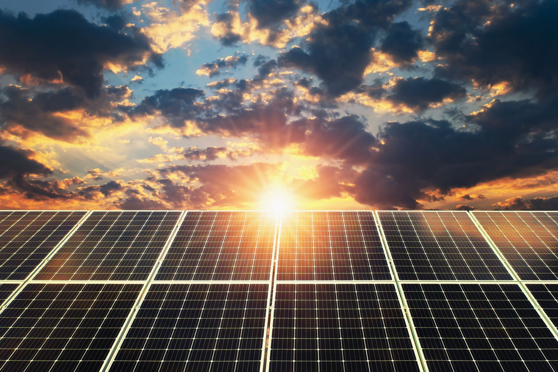 The sun is shining through the clouds over a field of solar panels.