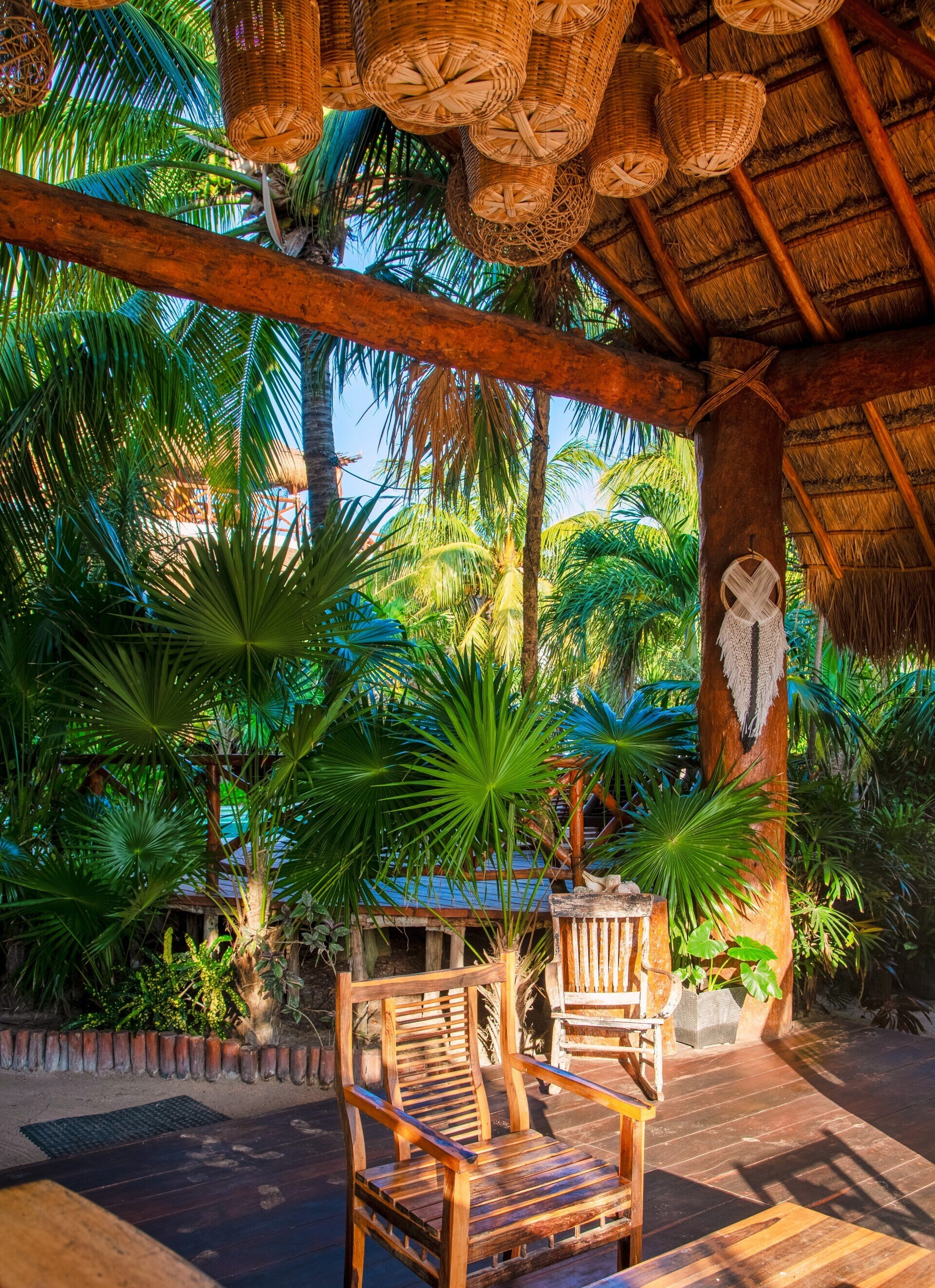 A patio with chairs and a table under a thatched roof surrounded by palm trees.