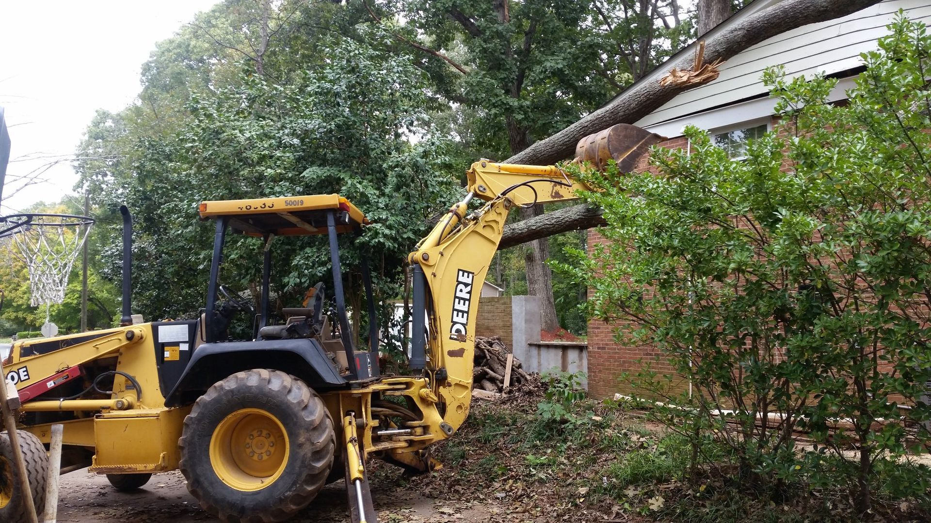 A yellow tractor is cutting down a tree in front of a house.