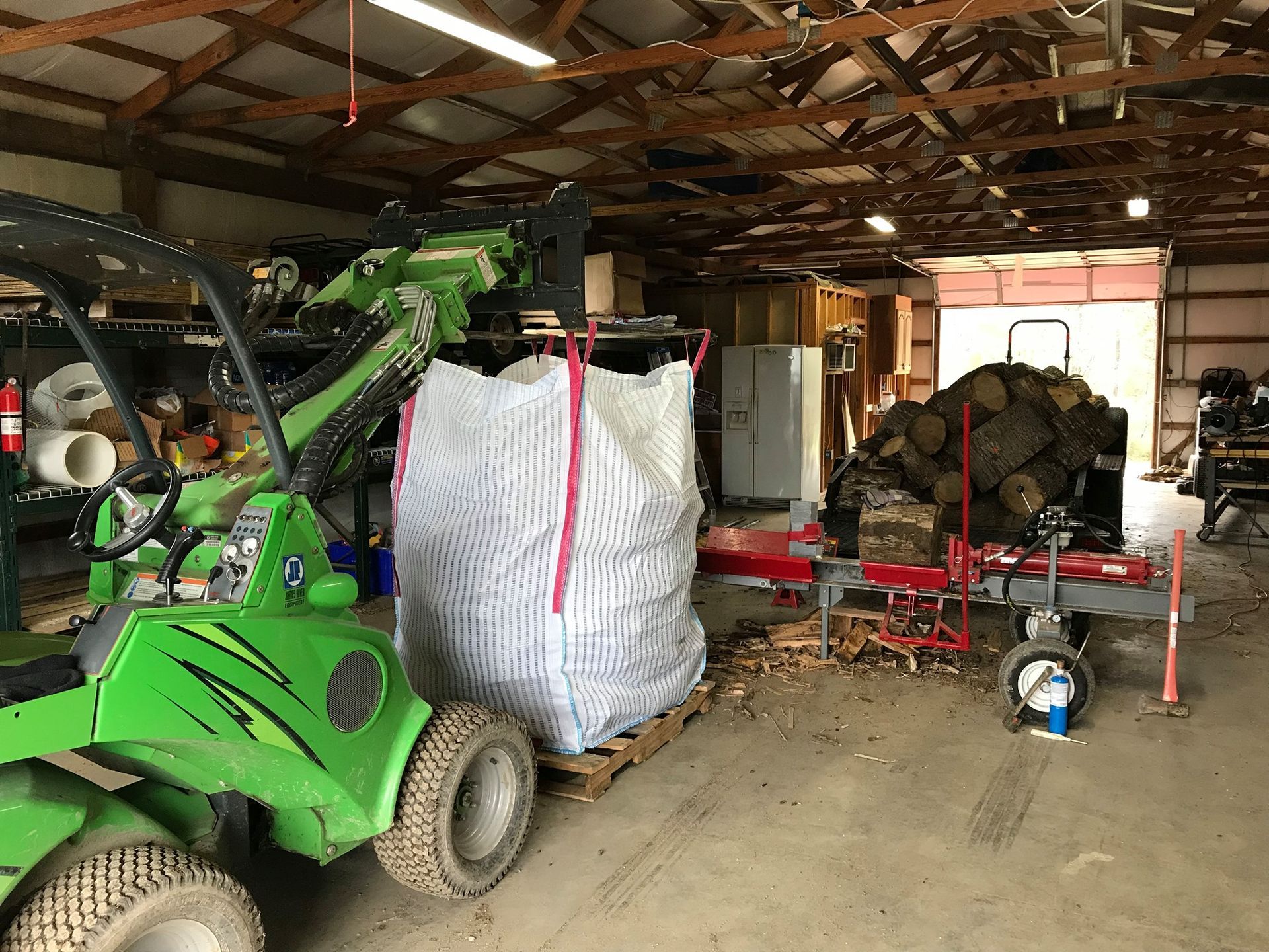 A green tractor is parked in a garage next to a pile of logs.