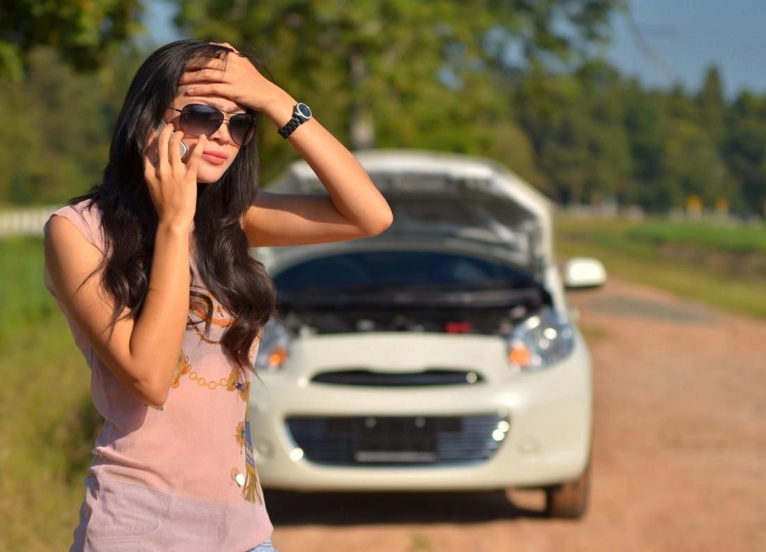 A woman is talking on a cell phone in front of a broken down car.