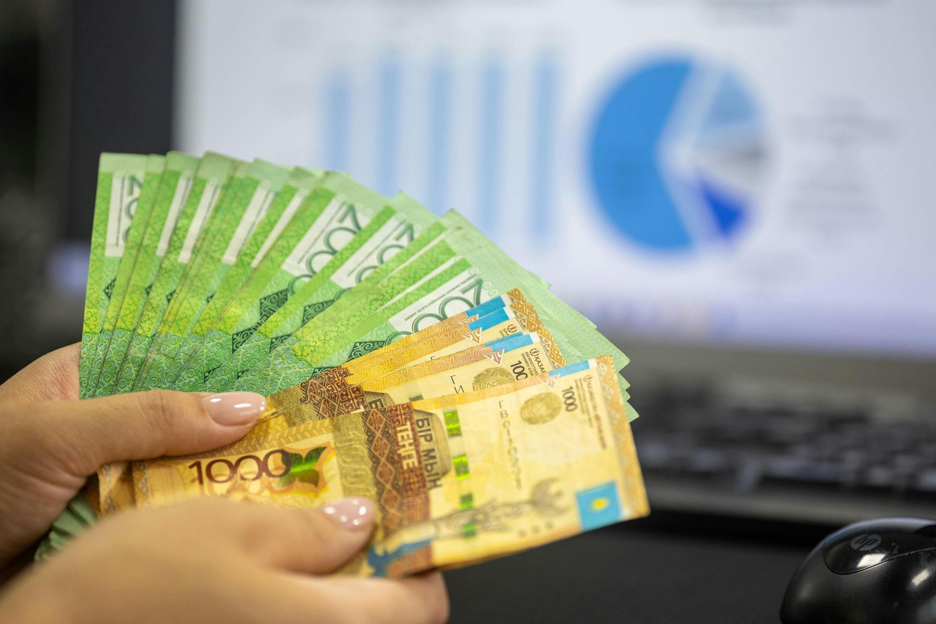 A person is holding a fan of money in front of a computer.
