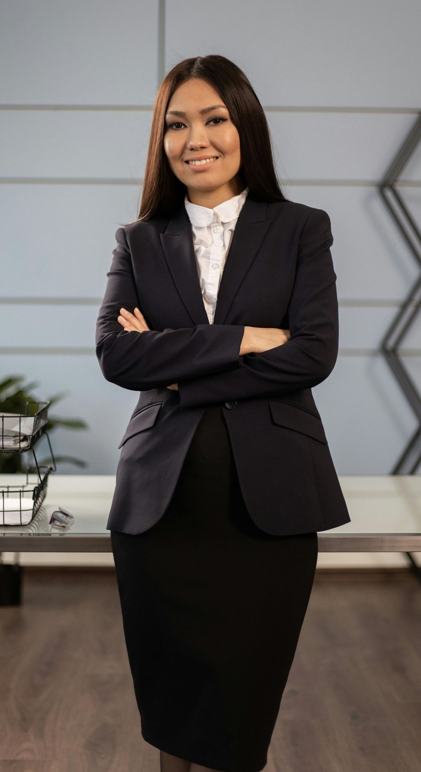 A woman in a business suit is standing in front of a desk with her arms crossed.