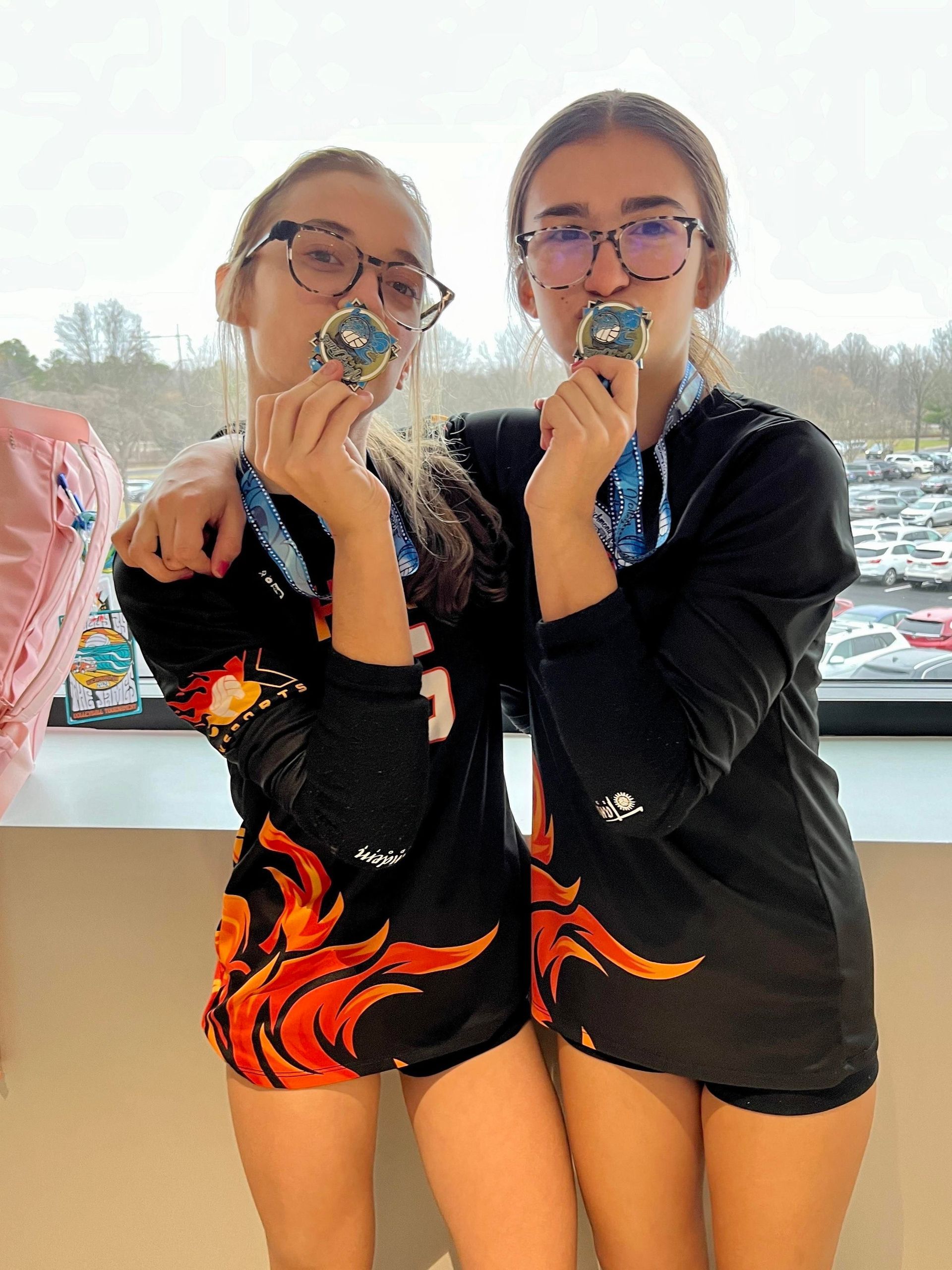 Two girls are standing next to each other holding medals in their mouths.