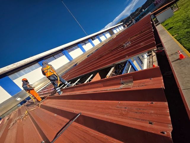 Dos trabajadores, equipados con trajes de seguridad y arneses, instalan paneles metálicos rojos en el techo de un edificio