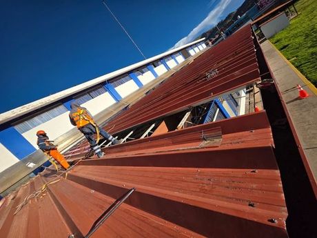 Dos trabajadores, equipados con trajes de seguridad y arneses, instalan paneles metálicos rojos en el techo de un edificio