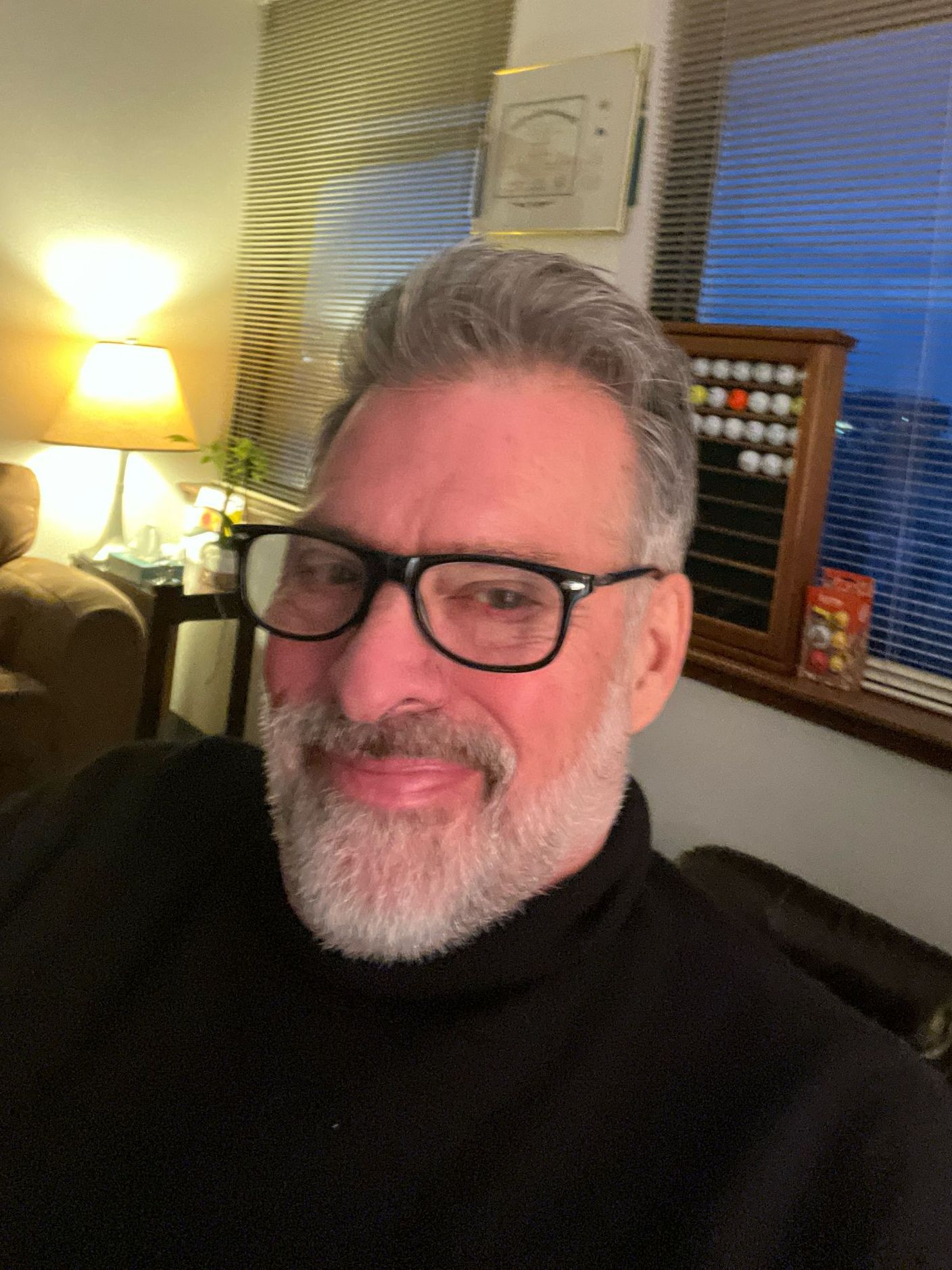 A man with glasses and a beard is sitting on a couch in a living room.