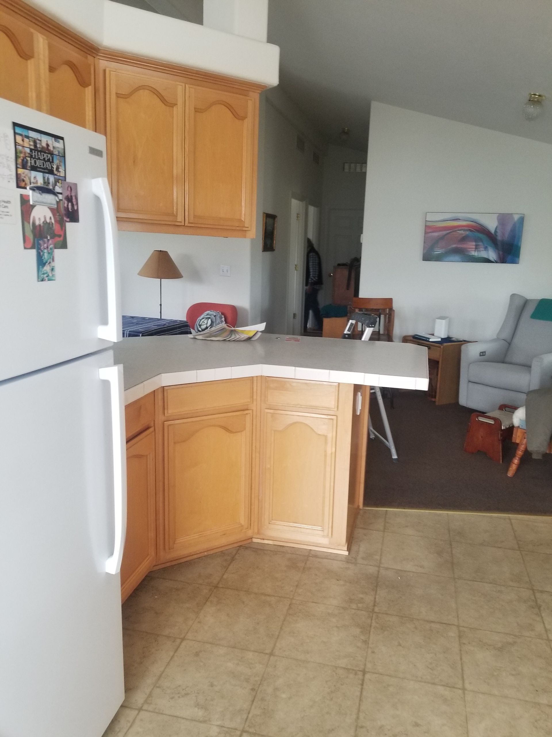 Kitchen with light-colored cabinets, a white refrigerator, and a countertop. A living area is visible in the background.