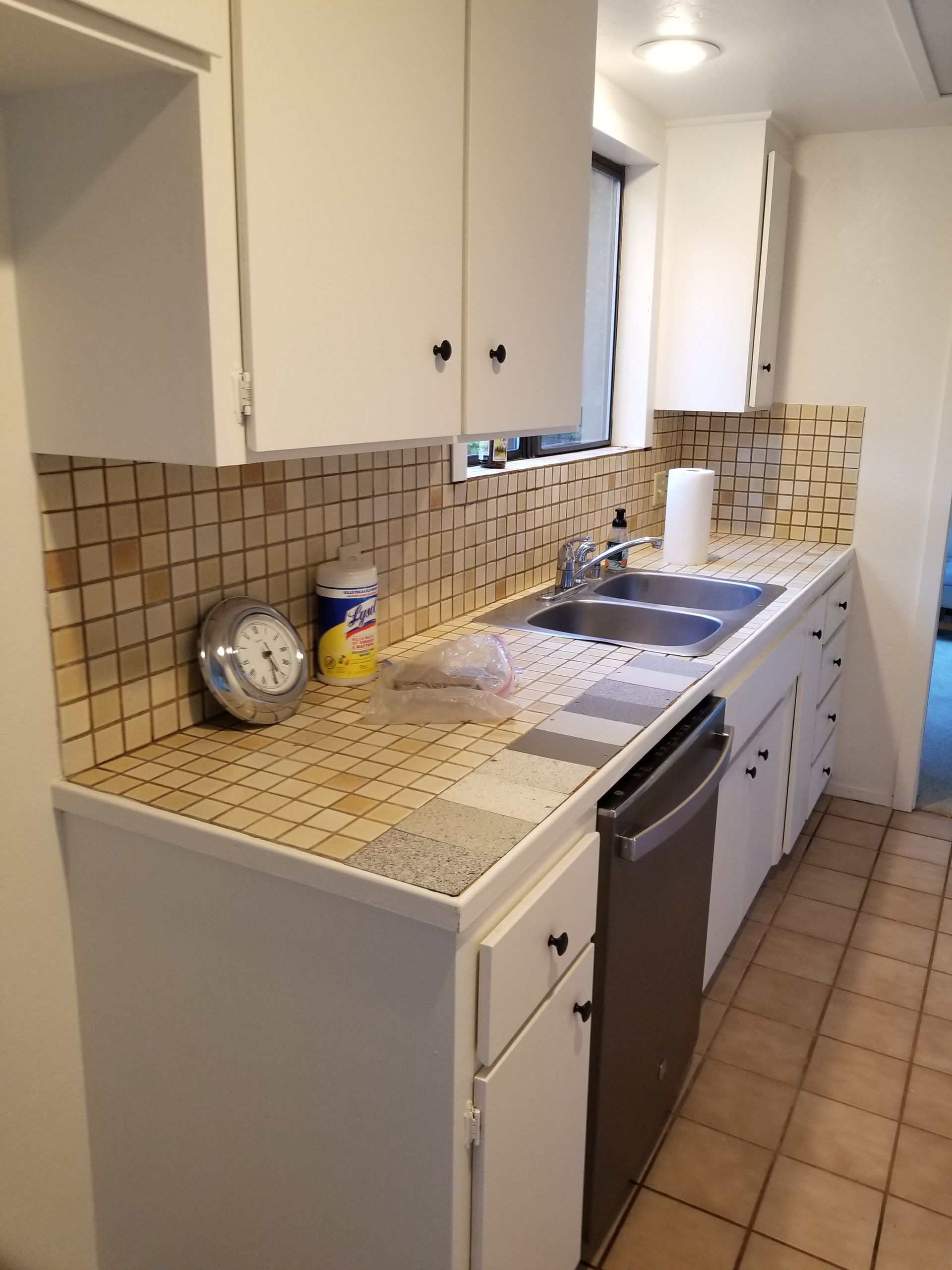 Kitchen with white cabinets, tan tile backsplash, stainless steel sink and dishwasher, and beige tile floor.
