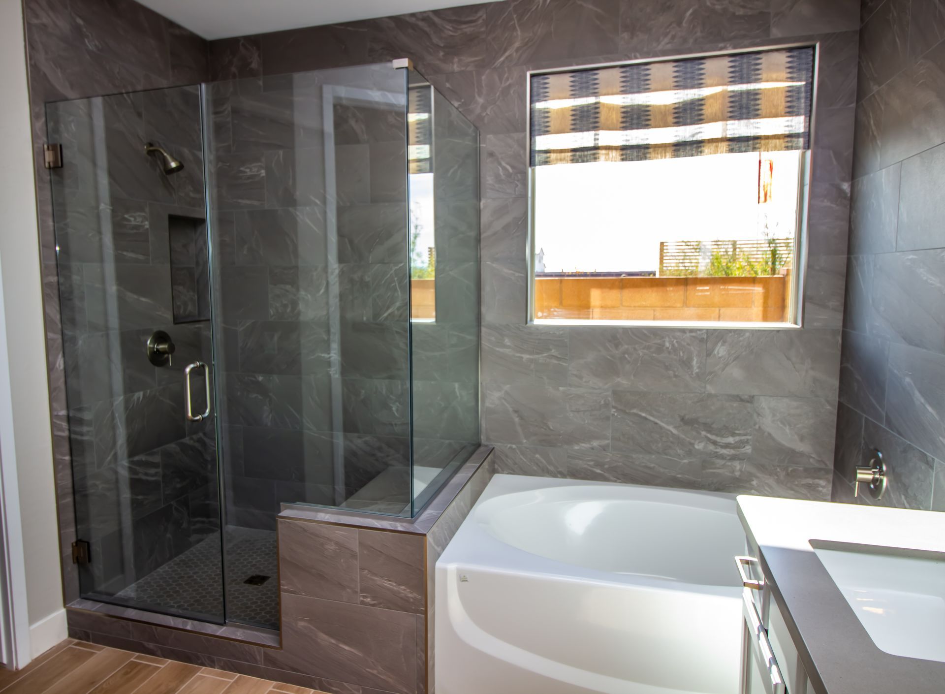 Modern bathroom with gray tile walls, glass shower, and a white bathtub.
