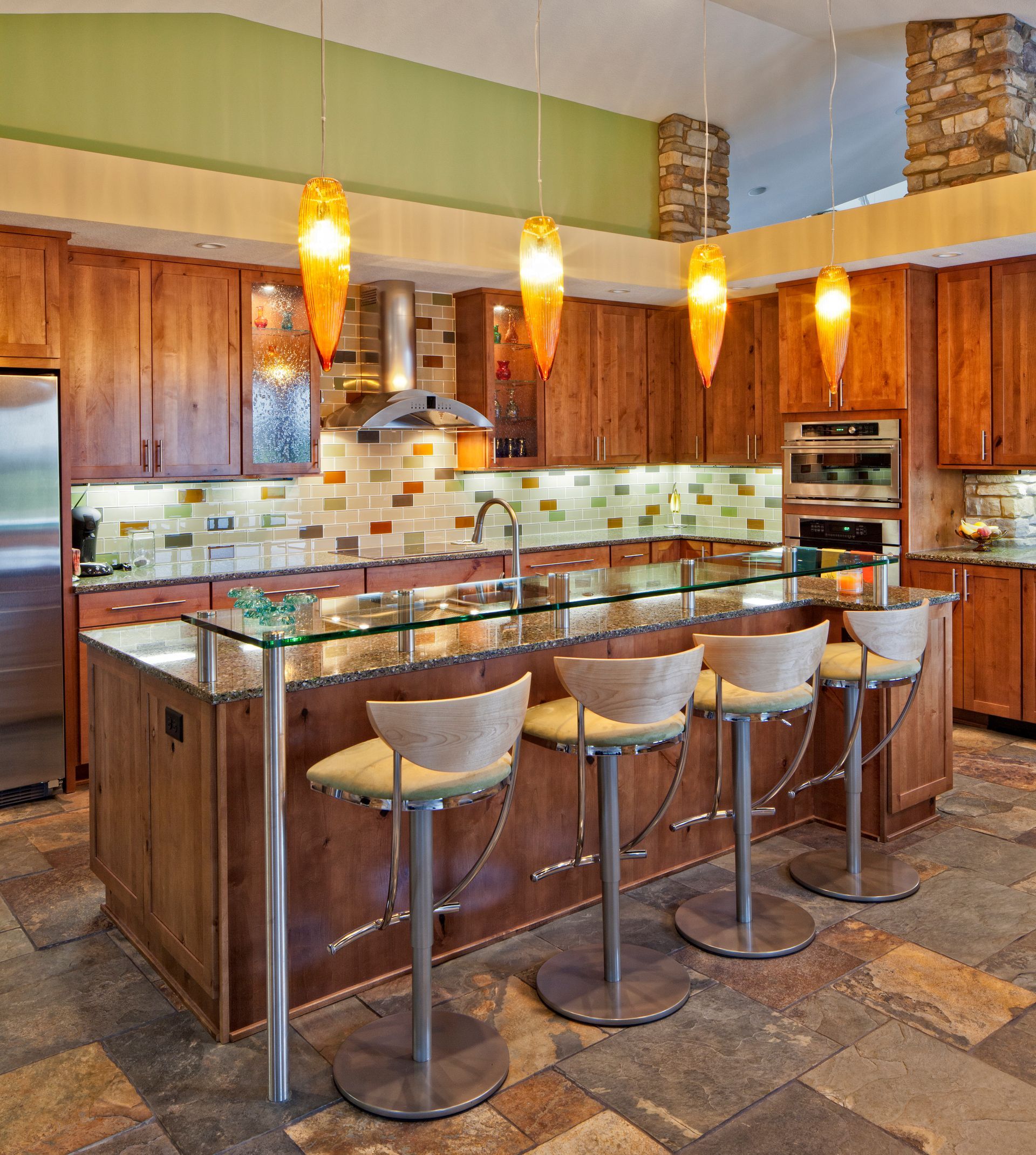 Modern kitchen with wooden cabinets, glass countertops, and bar stools; overhead orange pendant lights.