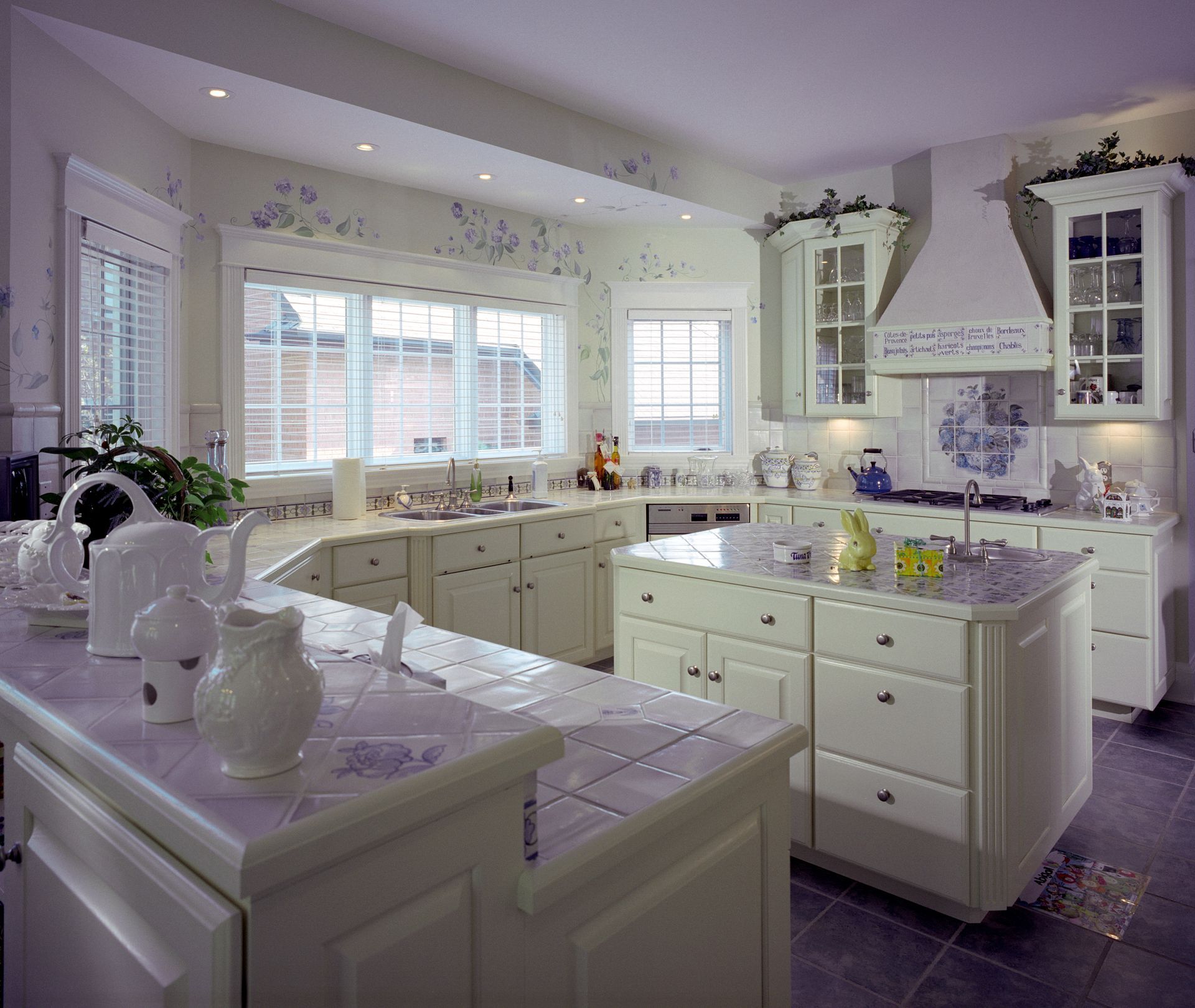 Bright, spacious kitchen with white cabinets, counters, and an island. Windows, cooktop, and appliances are also visible.