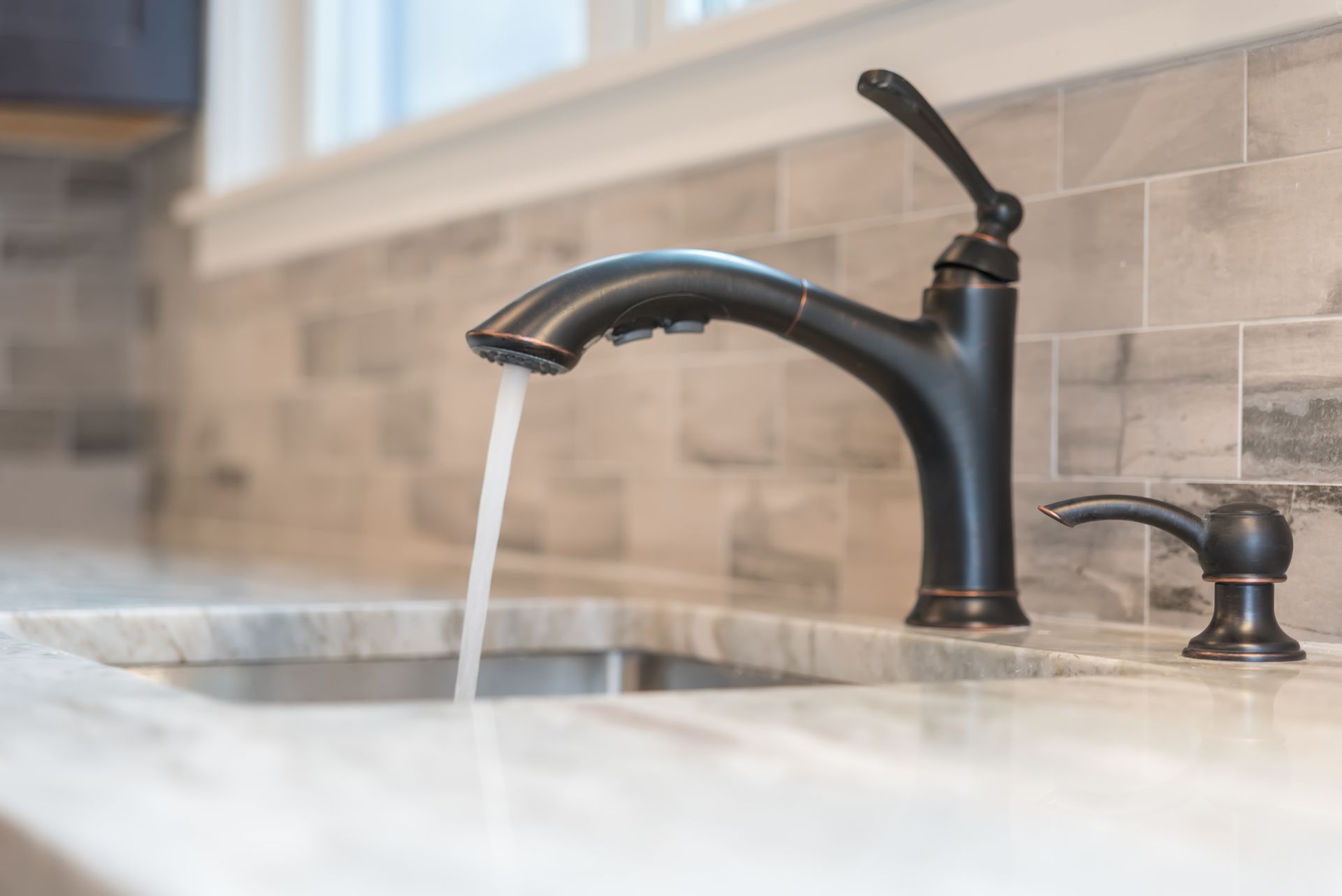Black faucet with running water in a kitchen sink; tile backsplash, countertop, and soap dispenser.
