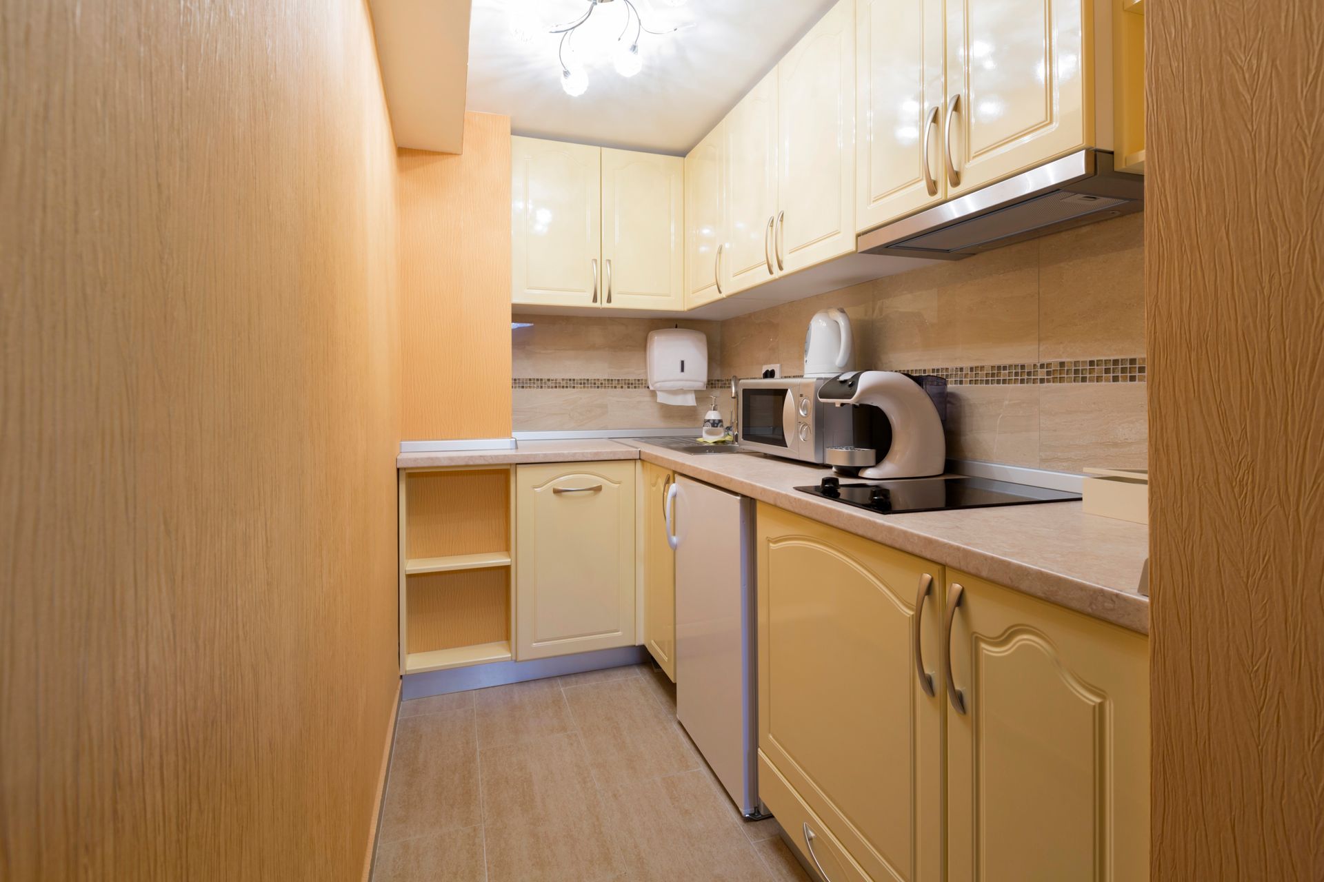 Small, pale yellow kitchen with cabinets, appliances, and light-colored tiled backsplash and a fridge.