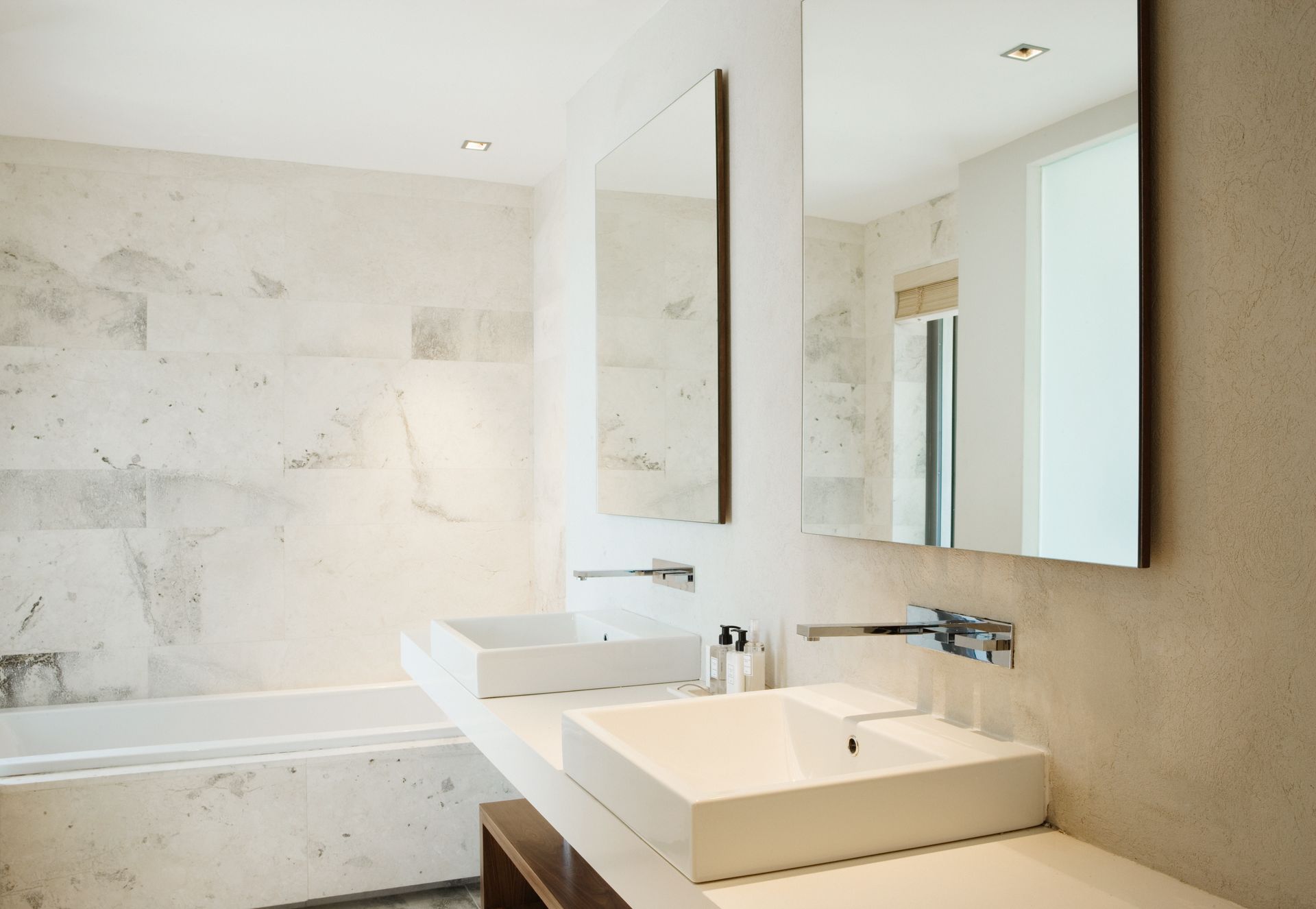 Modern bathroom with white sinks, marble wall, large mirror, and bathtub.
