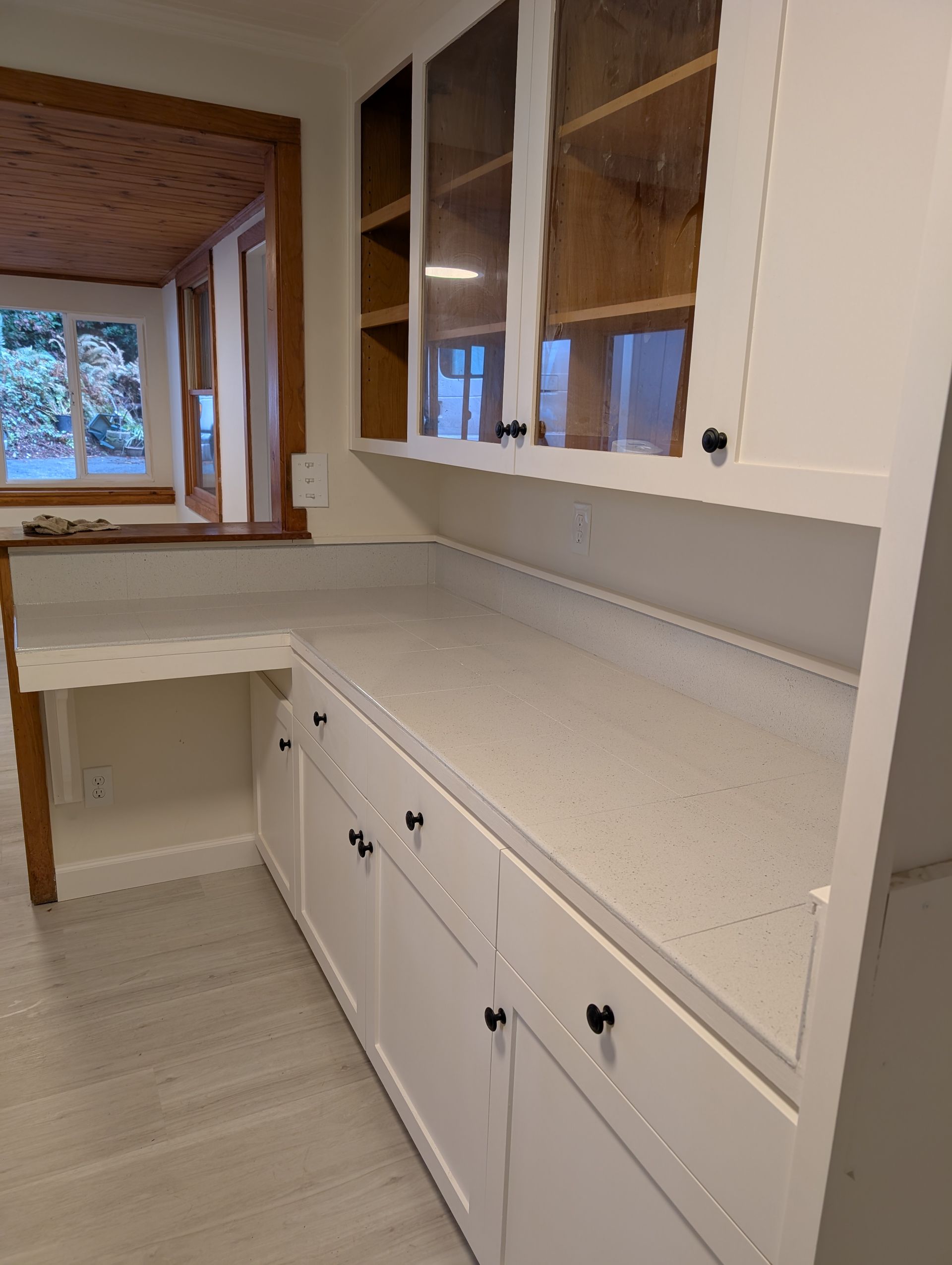 White kitchen cabinets with countertop and glass-door upper cabinets; window in background.