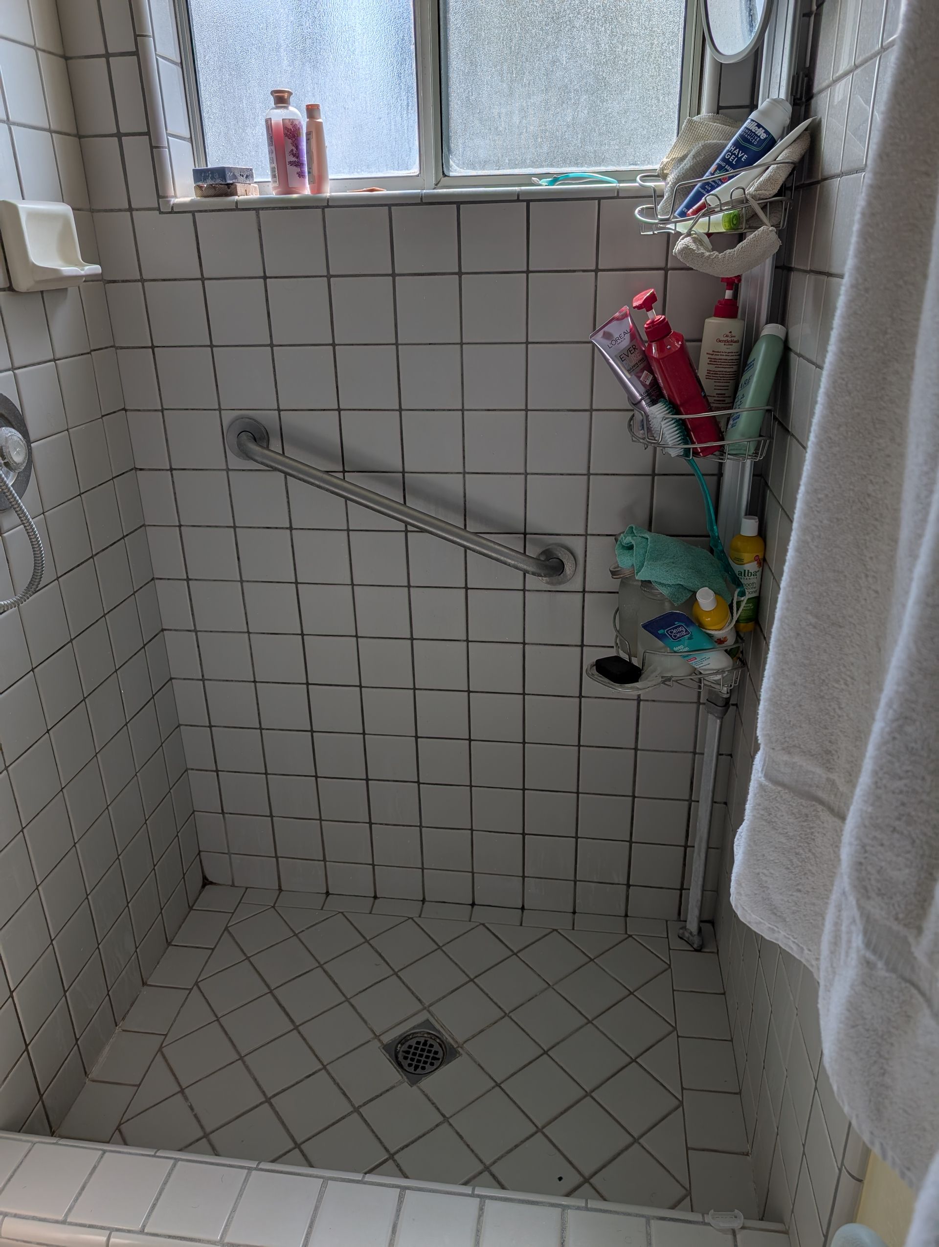 Small white tiled shower with a window, a grab bar, and toiletries.