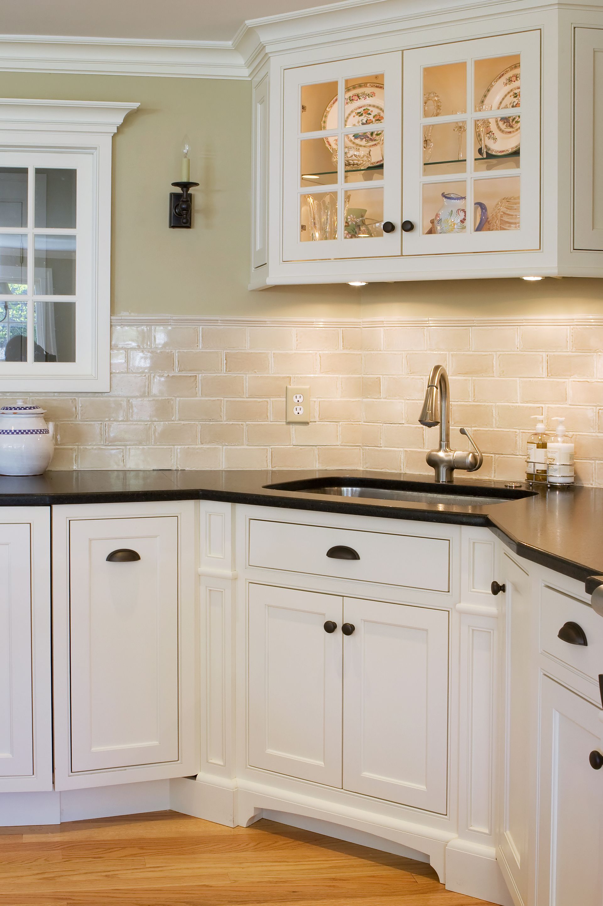 White kitchen cabinets with black countertop, light-colored backsplash, sink, and overhead glass-doored cabinets.