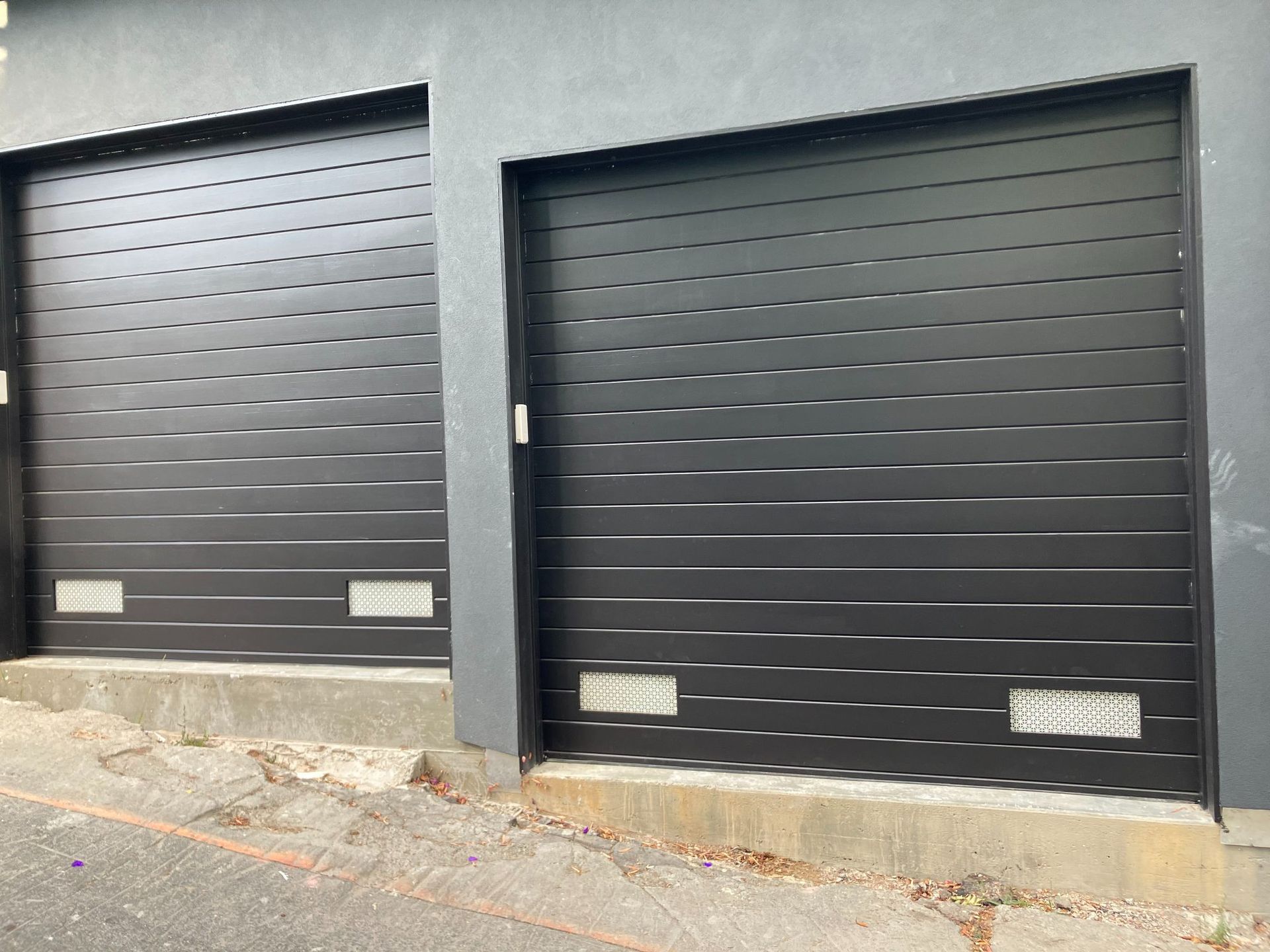 Two black garage doors on a gray building exterior with concrete pavement in front
