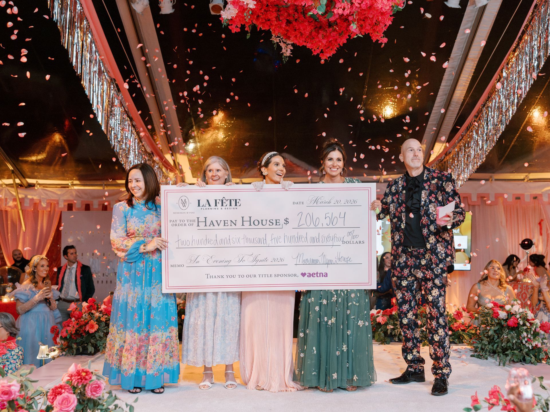 Four women in colorful outfits hold a large check at a festive nighttime celebration of An Evening To Ignite at Merrimon-Wynne in Raleigh, NC.