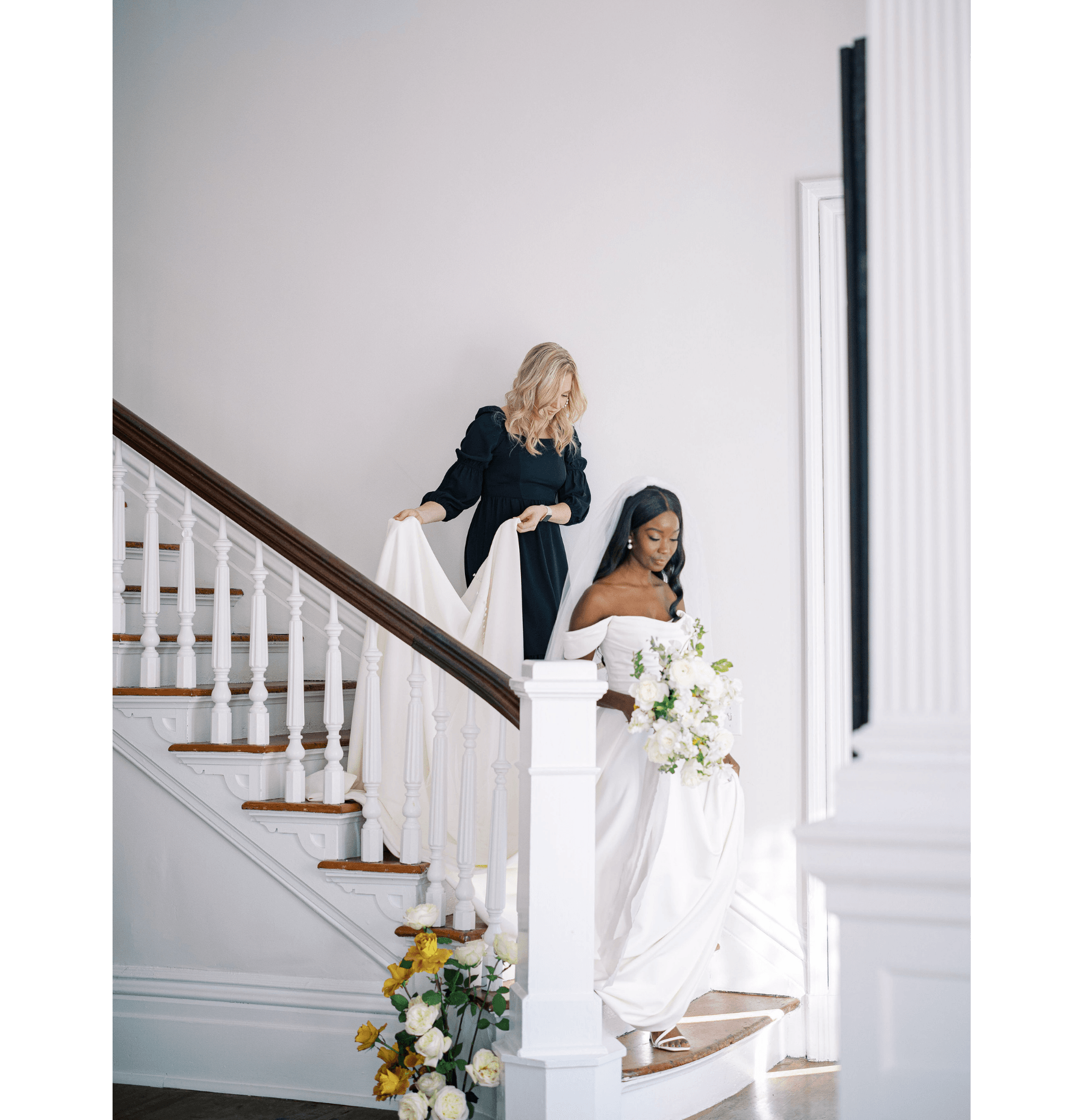 A wedding planner is helping a bride walk down a large staircase by holding her train of her wedding dress.