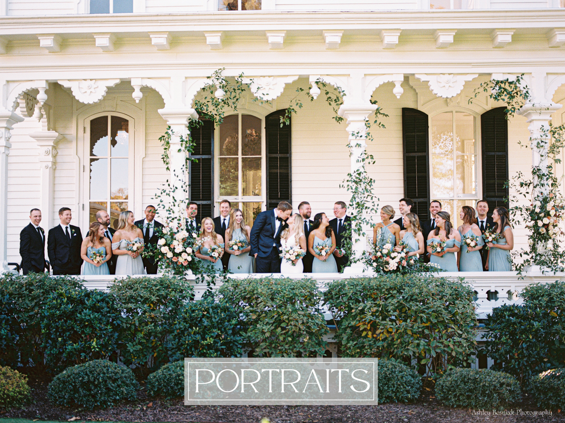 A large wedding party is standing on the large white porch of a house with flowers cascading up the columns of the porch.