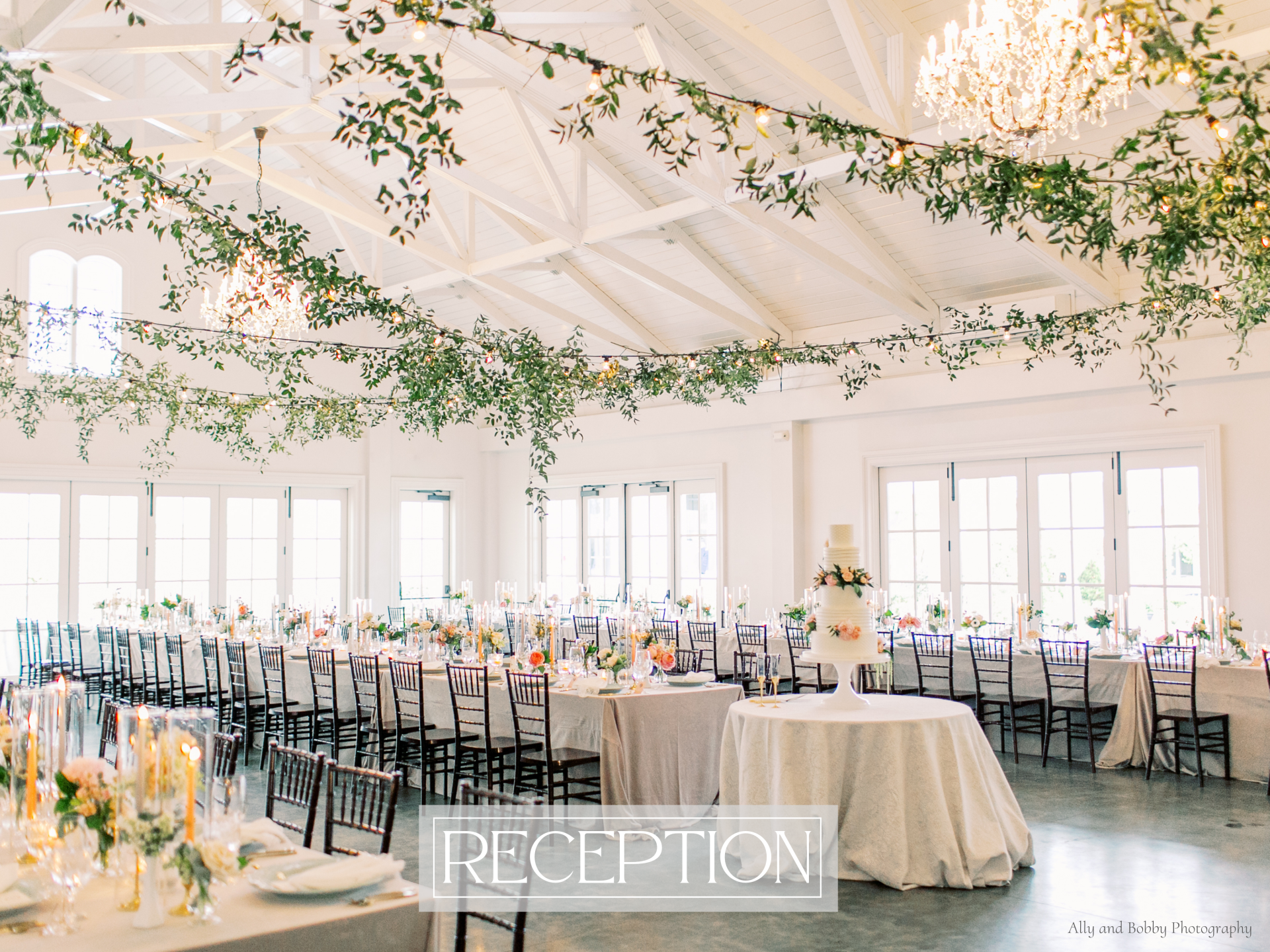 A large wedding ballroom with vaulted ceilings is full of tables and chairs set up for a wedding reception. There are vines of greenery drapping throughout the ceiling.