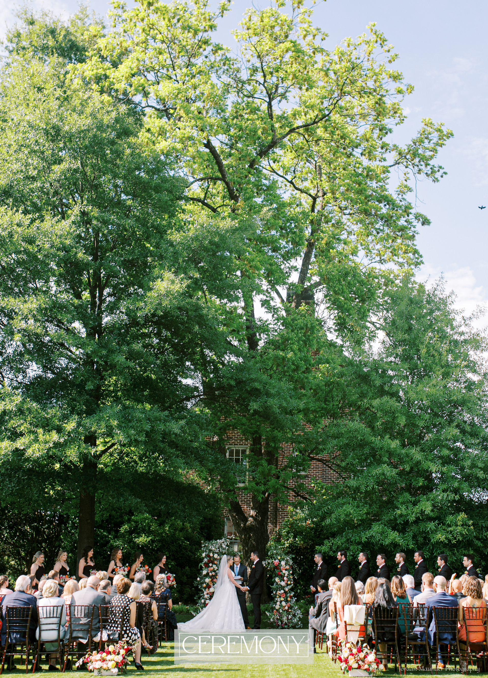 A bride and groom are getting married in front of a large full green oak tree with their bridal party members standing to their left and right and guests are sitting in rows of chairs.