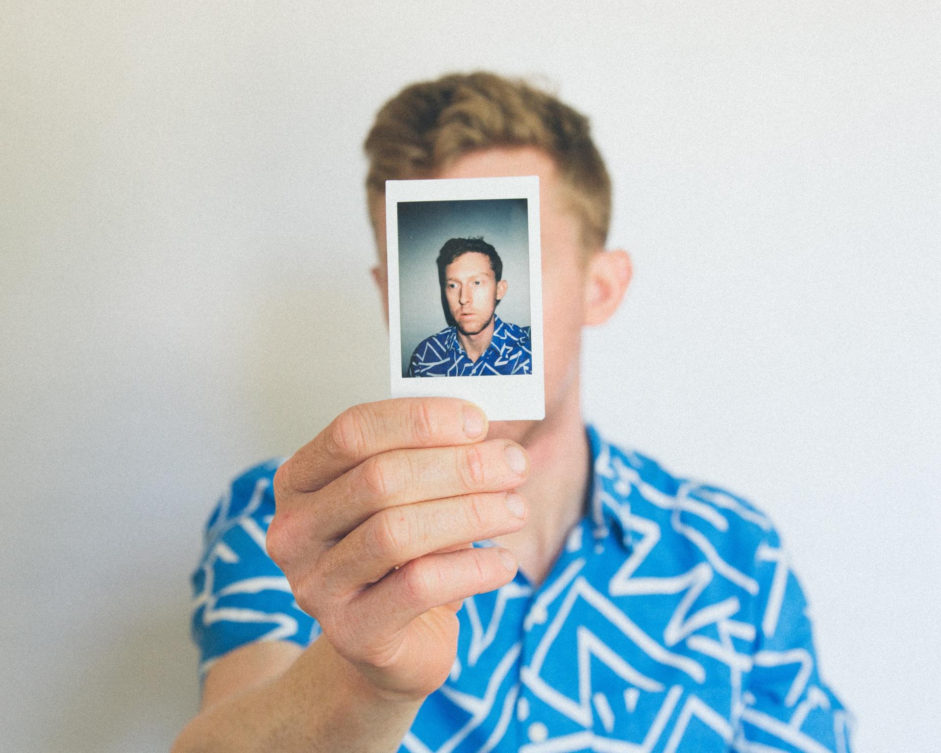 Man holding a Polaroid photo in front of his face. He wears a blue patterned shirt.