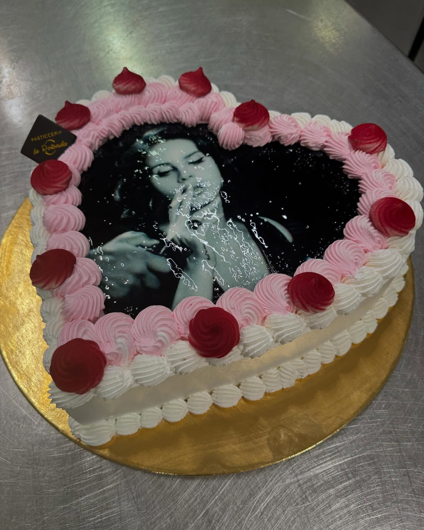 Torta a forma di cuore con la foto di una donna al centro, circondata da glassa rosa e bianca e guarnita con decorazioni rosse.