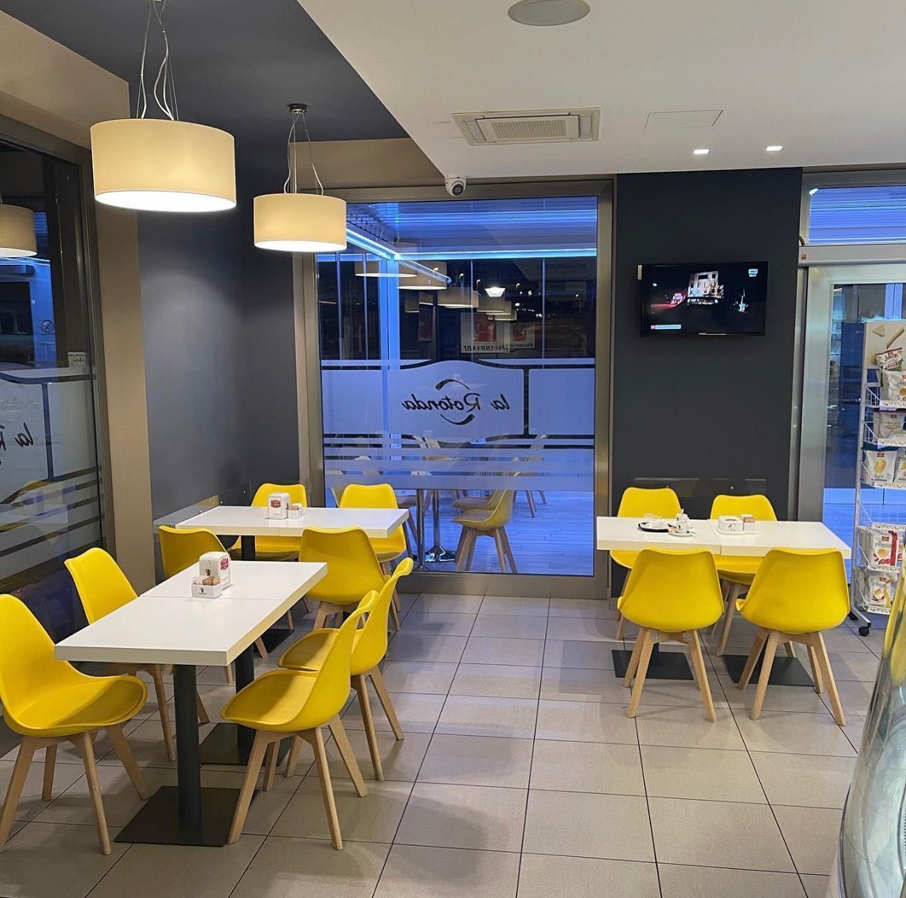 Interno di un bar con tavoli bianchi, sedie gialle e luci soffuse; una finestra si affaccia sulla strada.