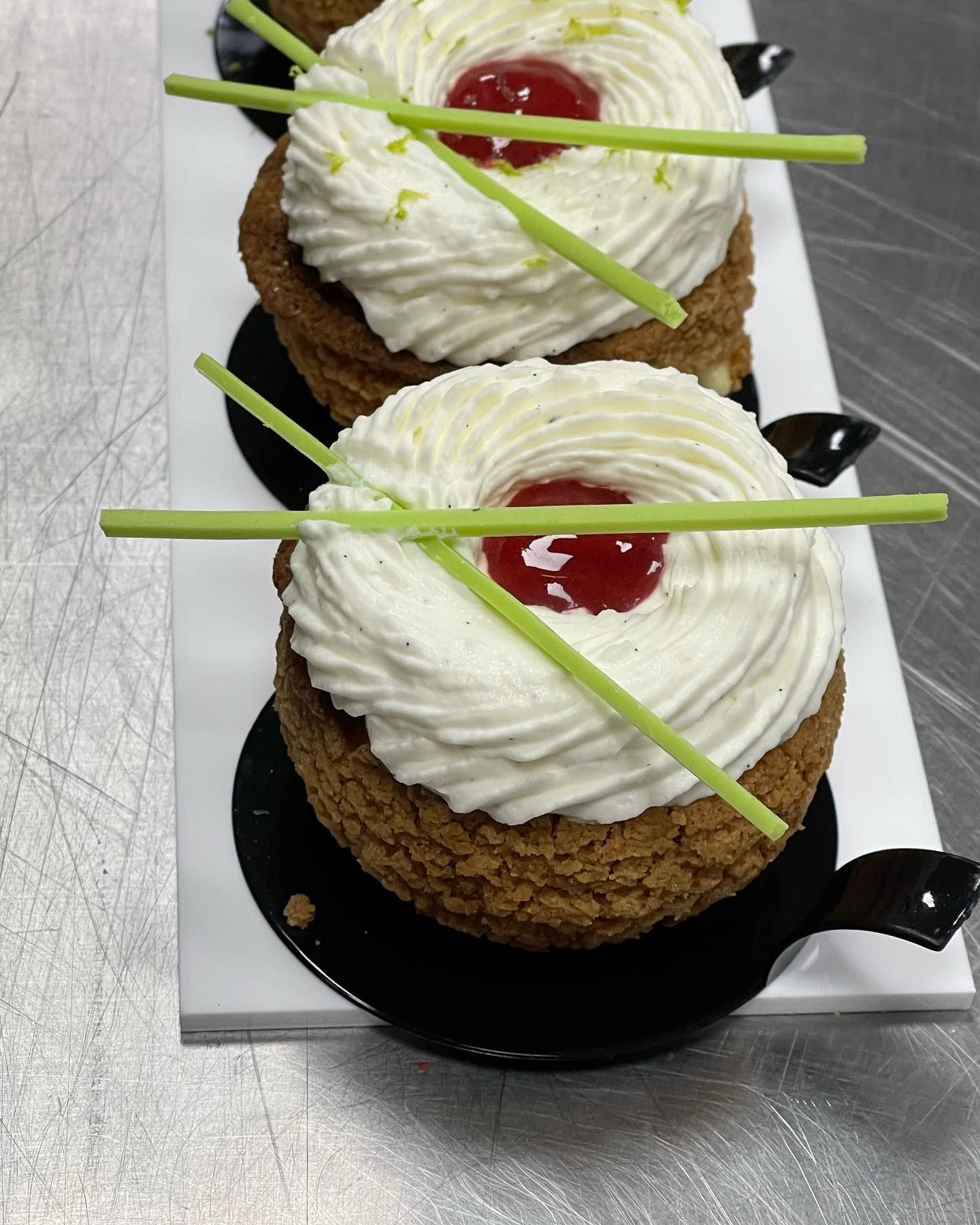 Due pasticcini ricoperti di crema, ciliegie, scorza di lime e bastoncini verdi decorativi su un vassoio bianco.