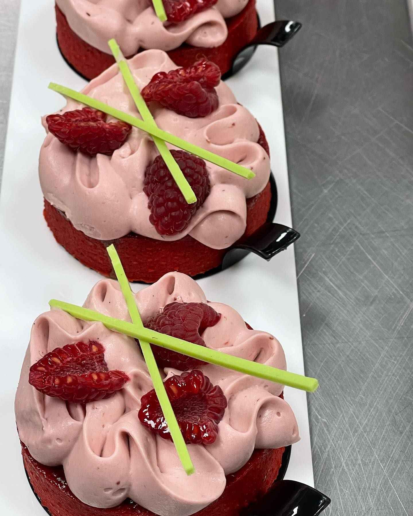 Tre crostatine ai lamponi con glassa rosa, lamponi freschi e cannucce di zucchero verdi su un vassoio bianco.