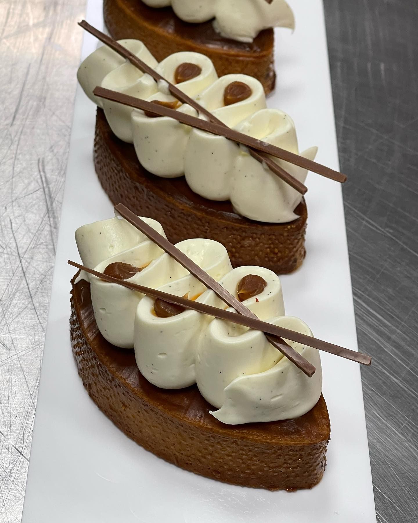 Tre tortine ovali al caramello, guarnite con panna montata bianca e bastoncini di cioccolato, su un piatto bianco.