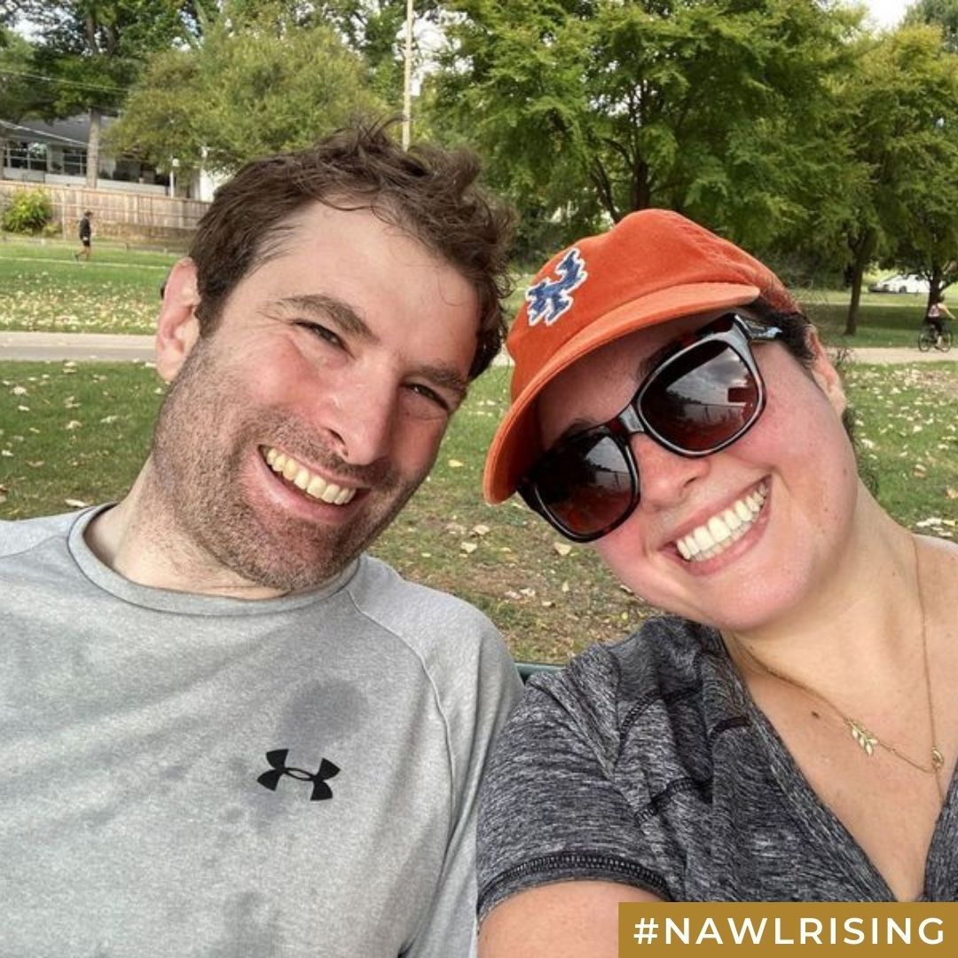 Two smiling people lean their heads together and smile while sitting on a grassy field.