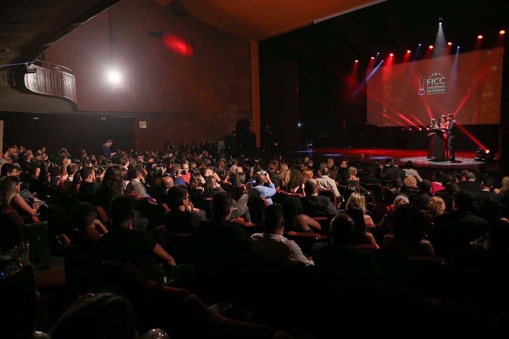 Uma grande multidão de pessoas está sentada em um teatro assistindo a uma apresentação.