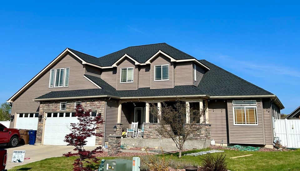 A large two-story house with a dark shingled roof, brown siding, and a white garage under a clear blue sky.