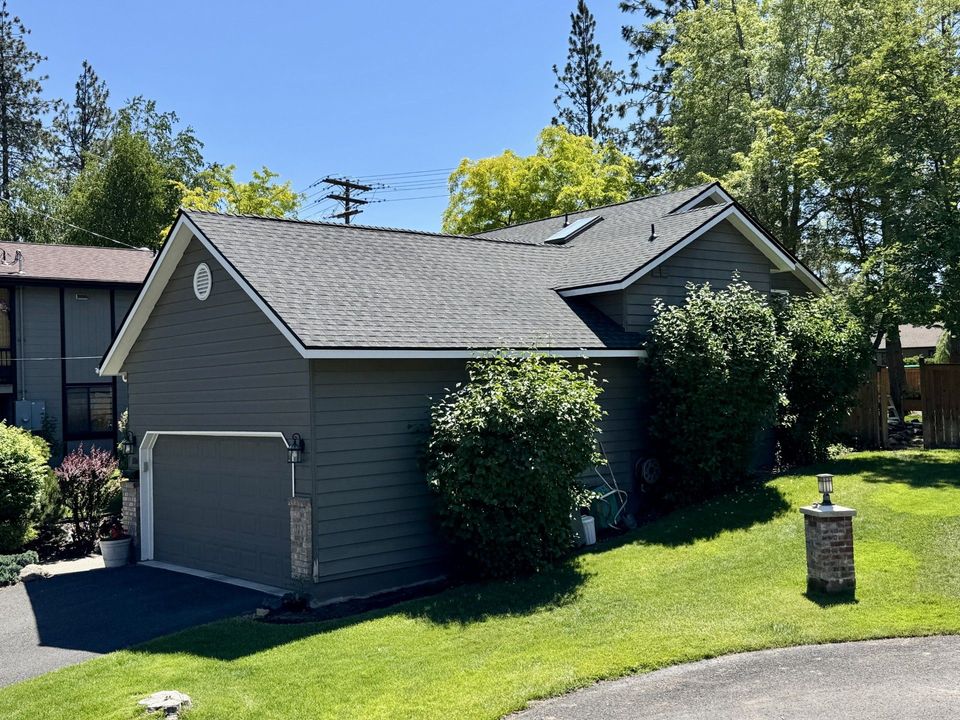 Gray house with a dark shingled roof, attached garage, and surrounding green lawn on a sunny day.