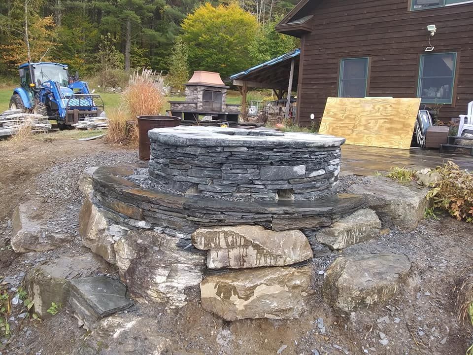 Stone fire pit being built outdoors. Blue tractor in the background near a house.